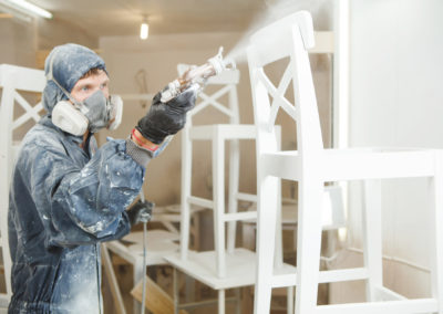 Man painting chair into white paint in respiratory mask. Application of flame retardant ensuring fire protection, airless spraying.